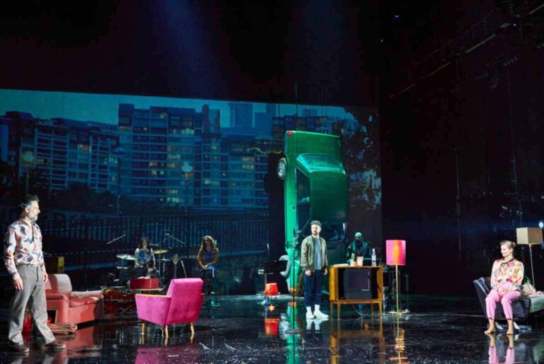 ARCHIV DER SEHNSÜCHTE, Mehrere Darsteller:innen stehen auf einer Theaterbühne um einen Fernseher und eine Lampe, während hinten ein Auto von der Decke hängt.