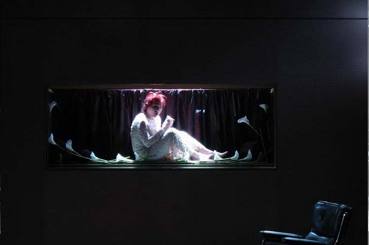 PRINZESSIN NICOLETTA Neumarkt Theater Zürich Bühne und Kostüme: Alissa Kolbusch. Eine Frau sitzt in einem weißen Kleid in einem Schaufenster hinter Glas mit Totenblumen.