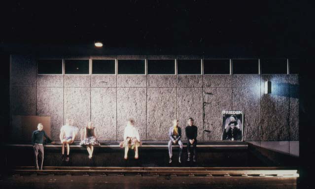 DER GETEILTE HIMMEL Volksbühne Berlin, Bühne Alissa Kolbusch. 2 Schauspieler stehen vor einem geöffneten Werks- Tor , 6 Schauspieler sitzen auf der Kante eines Bahnsteigs vor einer DDR- Plattenbau- Wand.