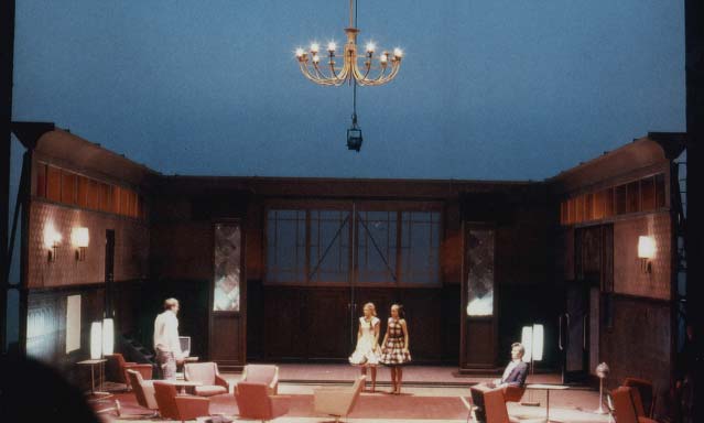 DER GETEILTE HIMMEL Volksbühne Berlin, Bühne Alissa Kolbusch. Ein rötlich-brauner DDR- Salon mit hinten 2 Säulen und vielen 60er Jahre DDR_Cocktail- Sesseln und Stehlampen. Darin stehen 2 Damen in Cocktailkleidern und schauen nach links vorne zu einem Mann. Ein anderer Mann sitzt in einem Sessel.