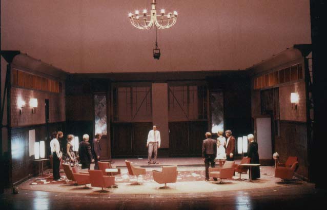 DER GETEILTE HIMMEL Volksbühne Berlin, Bühne Alissa Kolbusch. Ein rötlich-brauner DDR- Salon mit hinten 2 Säulen und vielen 60er Jahre DDR_Cocktail- Sesseln und Stehlampen. Darin stehen ca. 10 Schauspieler in Anzügen und Schauspielerinnen in Cocktailkleidern um eine Grube herum.
