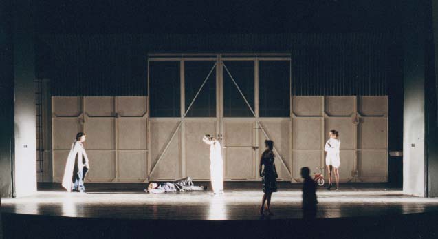 DER GETEILTE HIMMEL Volksbühne Berlin, Bühne Alissa Kolbusch. 4 Schauspieler stehn vor einem WErks- Tor , einer liegt davor auf dem Boden.
