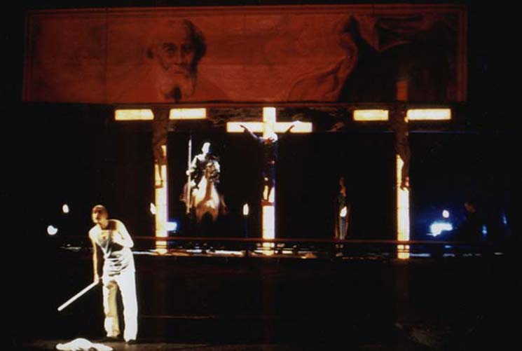 GOLDBERGVARIATIONEN, Theater Osnabrück, Ausstattung Alissa Kolbusch. Drei beleuchtete Kreuze, dahinter ein Reiter auf einem echten Pferd. Vorne steht ein Mann mit einem Stock in der Hand.