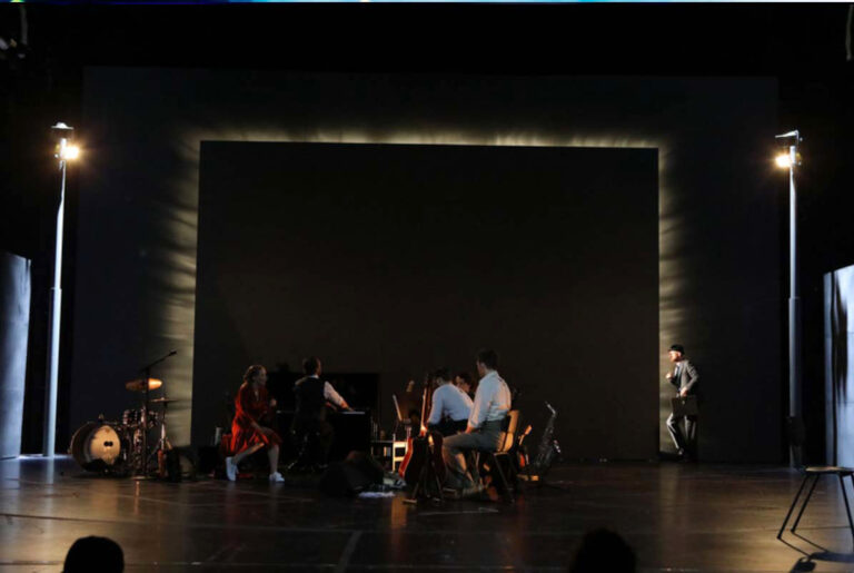 DER REISENDE,Theaterszene mit mehreren Darsteller:innen und Musikern in einer großen Wand, wo aus Schlitzen starkes Licht hervorkommt. rechts und libks 2 Flutlicht- Laternen.
