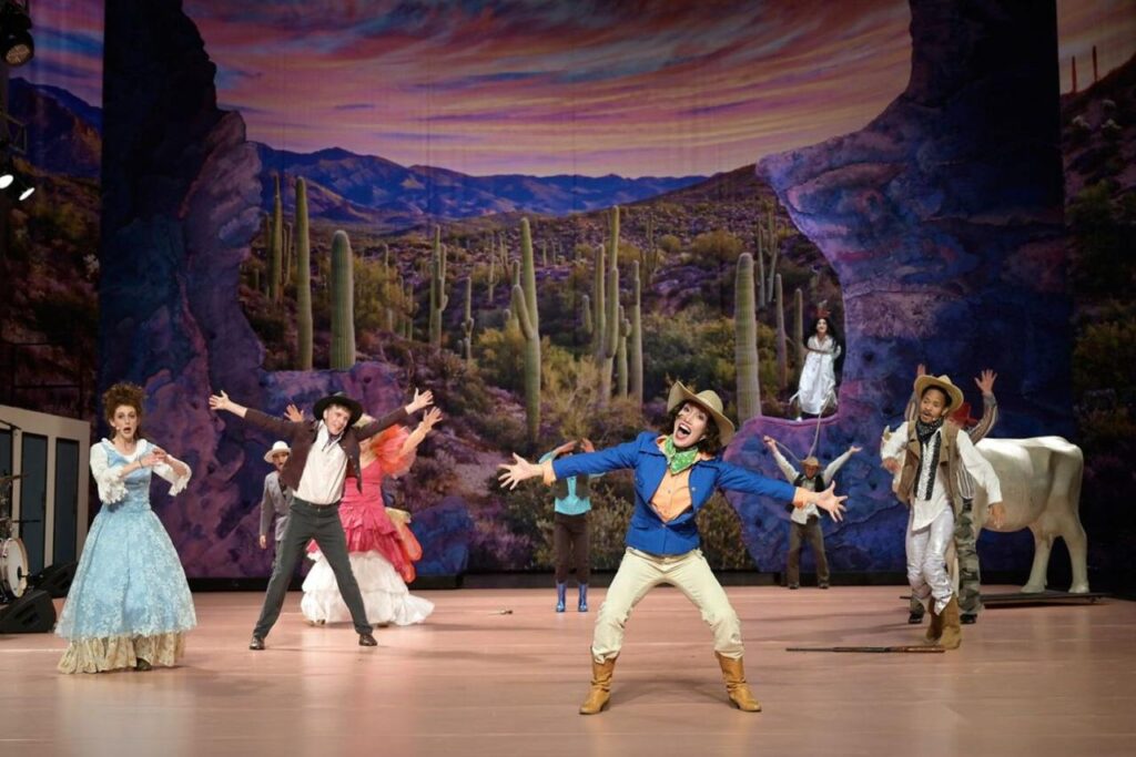 THE WEST, Volksbühne Berlin. Bühne: Alissa Kolbusch. Mehrere Darsteller:innen tanzen auf einer großen Theaterbühne mit Felsen vor einer Wüstenlandschaft.