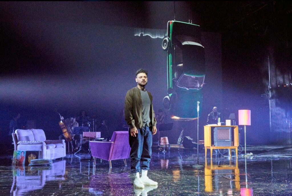ARCHIV DER SEHNSÜCHTE, Darsteller steht auf einer Theaterbühne zwischen Wohnzimmermöbeln und einem Fernseher, während hinten ein Auto von der Decke hängt.
