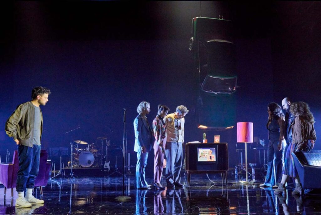 ARCHIV DER SEHNSÜCHTE, Mehrere Darsteller:innen stehen auf einer Theaterbühne um einen Fernseher und eine Lampe, während hinten ein Auto von der Decke hängt.