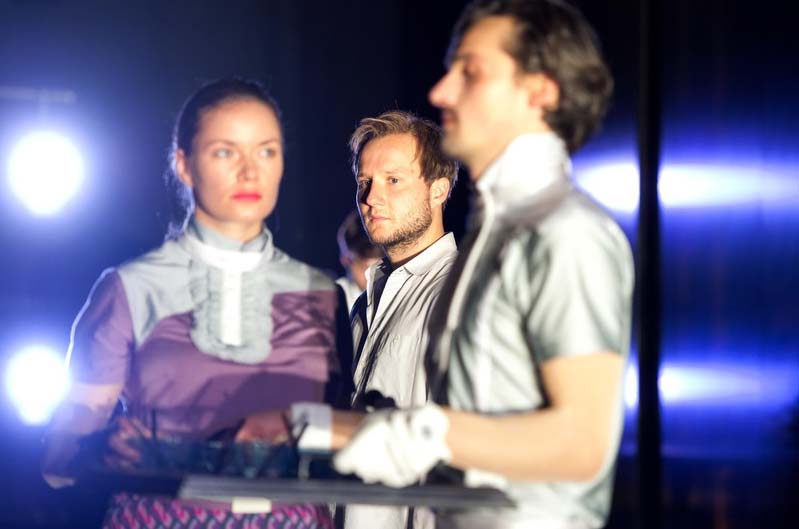BRAVE NEW WORLD Theater Bonn, Kostüme: Alissa Kolbusch. Eine Darstellerin mit einer lila Bluse schaut den Darsteller im grauen Kostüm an, der ein Tablett vor sich hält.