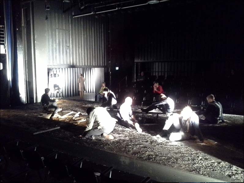 IM WESTEN NICHTS NEUES Staatstheater Braunschweig, Bühne und Kostüme: Alissa Kolbusch. Auf einer breiten Fläche in der Mitte sitzen zehn Darsteller als Soldaten des 1. Weltkriegs um ein Loch mit Holzkreuzen herum. Hinten kommt eine Frau durch ein Tor. Hinten und vorne sieht man Zuschauer- Reihen, oben viele Leuchtstofflampen.