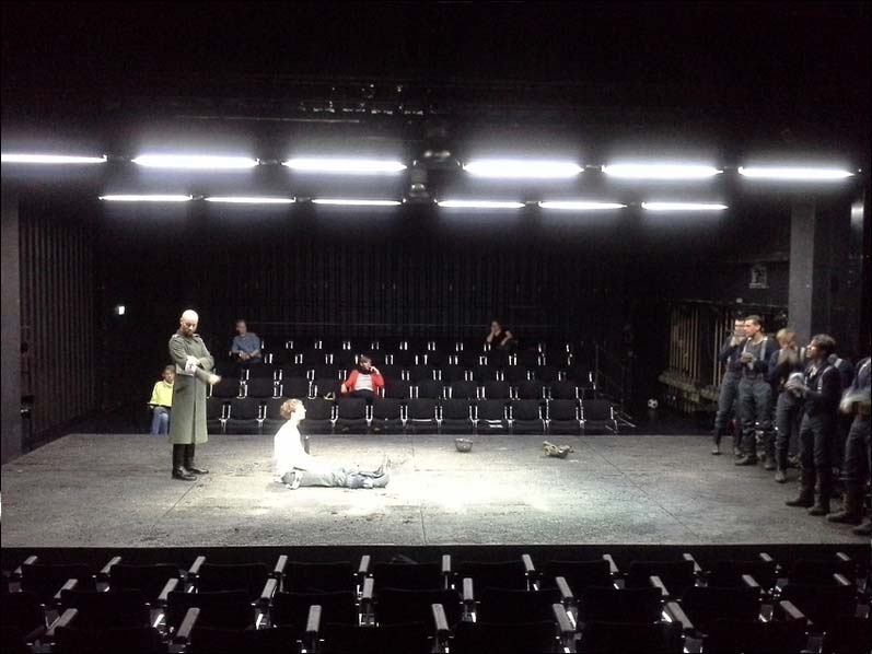 IM WESTEN NICHTS NEUES Staatstheater Braunschweig, Bühne und Kostüme: Alissa Kolbusch. Auf einer breiten Fläche in der Mitte stehen rechts acht Darsteller als Soldaten, in der Mitte sitzt einer auf dem Boden, hinter ihm steht ein Oberst im Mantel. Hinten und vorne sieht man Zuschauer- Reihen, oben viele Leuchtstofflampen.