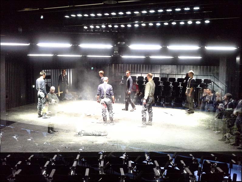 IM WESTEN NICHTS NEUES Staatstheater Braunschweig, Bühne und Kostüme: Alissa Kolbusch. Auf einer breiten Fläche in der Mitte laufen und stehen zehn Darsteller als Soldaten des 1. Weltkriegs um ein Loch herum. Hinten und vorne sieht man Zuschauer- Reihen, oben viele Leuchtstofflampen.