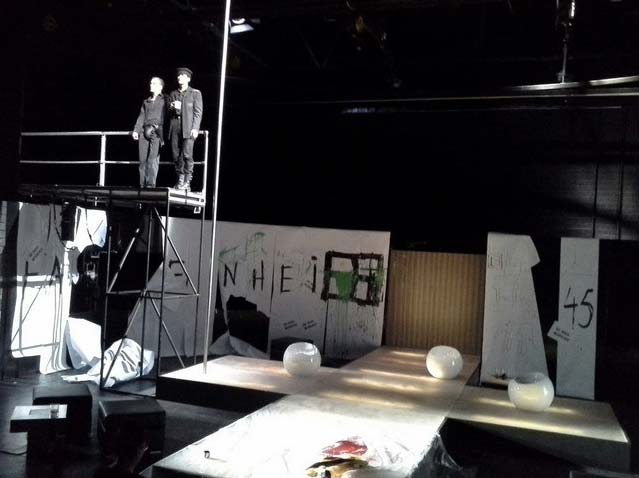 FAHRENHEIT 541 Hans- Otto- Theater Potsdam, Bühnenbild: Alissa Kolbusch. Ein weißes Kreuz aus Stegen mit 3 Sitzkugeln darauf, links oben auf einem hohen Podest stehen 2 Schauspieler nebeneinander und schauen nach vorne. Hinten weiße Papierbahnen auf den schwarzen Wänden.