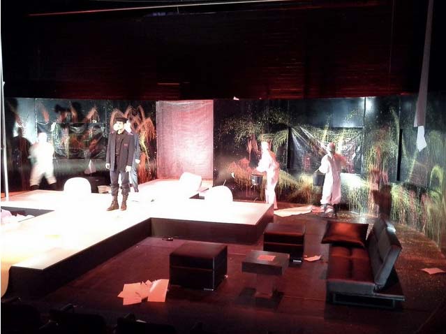 FAHRENHEIT 541 Hans- Otto- Theater Potsdam, Bühnenbild: Alissa Kolbusch. Ein Darsteller steht in der Mitte eines weißen gekreuzten Podestes, hinten malen Darsteller*innen in weißen Anzügen Feuer auf schwarze Wände. rechts vorne eine Sitzecke mit Sofa.