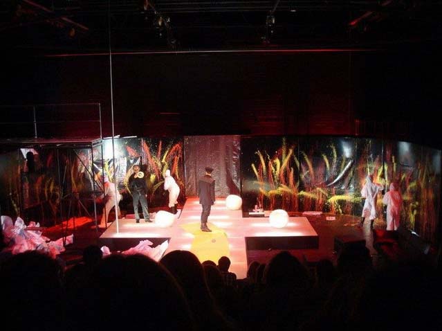 FAHRENHEIT 541 Hans- Otto- Theater Potsdam, Bühnenbild: Alissa Kolbusch. Ein Darsteller steht in der Mitte eines weißen gekreuzten Podestes, hinten malen Darsteller*innen in weißen Anzügen Feuer auf schwarze Wände.