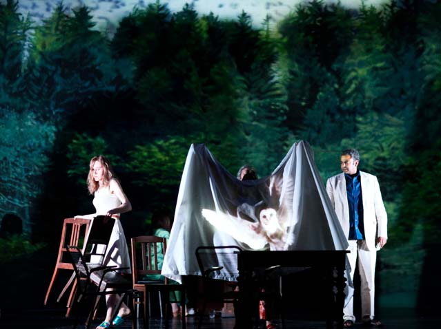 ONKEL WANJA Maxim Gorki Theater Berlin. Bühnenbild: Alissa Kolbusch. 1 Schauspielerin trägt zwei Stühle weg, 1 Schauspielerin hält eine Tischdecke hoch, auf die eine Eule projiziert wird. rechts daneben schaut ein Schauspieler in weißem Anzug zu.