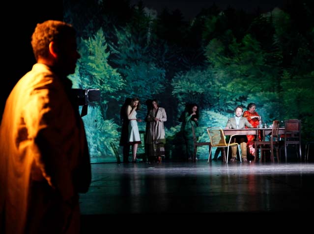 ONKEL WANJA Maxim Gorki Theater Berlin, Bühnenbild: Alissa Kolbusch. Vier Darstellerinnen stehen vor der Wand mit dem gemalten Wald, Ein Schauspieler sitzt hinten am Tisch. Im Vordergrund links steht ein andere Schauspieler. Hinten die Kulisse eines gemalten Waldes in einem Halbrund.