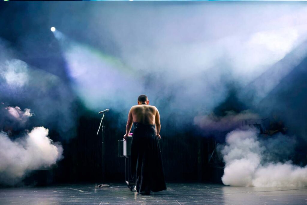UNSER DEUTSCHLANDMÄRCHEN, Darsteller steht mit dem Rücken zum Publikum auf einer nebelverhangenen Theaterbühne neben einem Mikrofon unter hellem Bühnenlicht.
