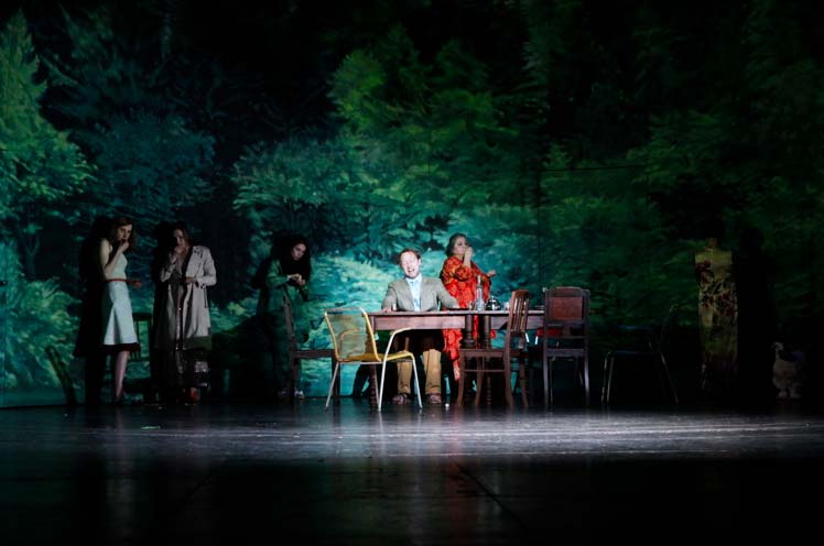 ONKEL WANJA Maxim Gorki Theater Berlin, Bühnenbild: Alissa Kolbusch. 4 Darstellerinnen stehen vor der Wand mit dem gemalten Wald, Ein Schauspieler sitzt am Tisch. Hinten die Kulisse eines gemalten Waldes in einem Halbrund.