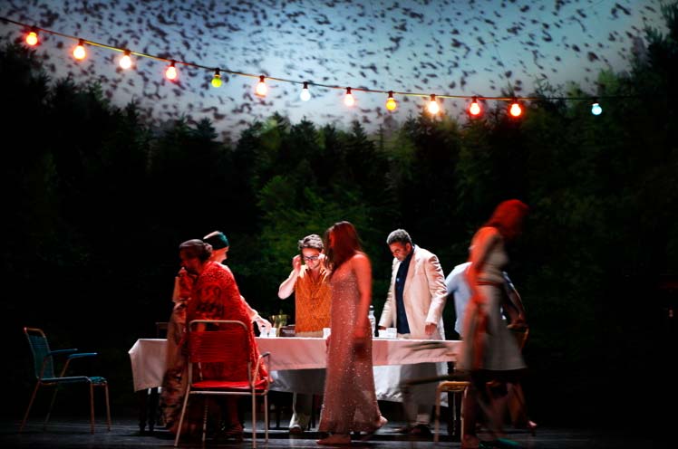 ONKEL WANJA Maxim Gorki Theater Berlin, Bühnenbild: Alissa Kolbusch. Sieben Darsteller laufen um einen Tisch herum, oben hängt eine bunte Lichterkette, hinten die Kulisse eines gemalten Waldes in einem Halbrund mit aufprojiziertem Vogelschwarm.