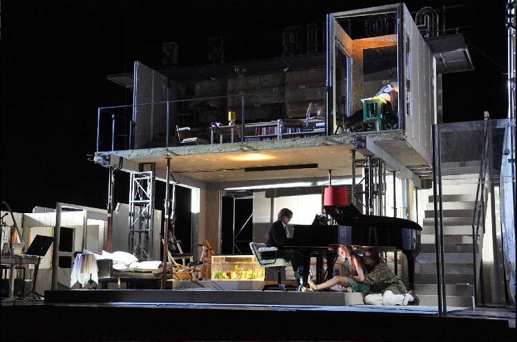 RHEINGOLD, Staatsschauspiel Dresden, Bühne und Kostüme Alissa Kolbusch. ein 2- stöckiges Rohbau- Haus, provisorisch eingerichtet, mit einer Treppe, Geländer und Möbeln. Unten ein Pianist am Flügel und 2 Darsteller*innen, die singend unter dem Flügel sitzen.