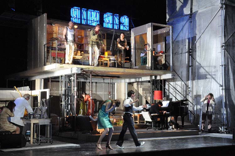 RHEINGOLD, Staatsschauspiel Dresden, Bühne und Kostüme Alissa Kolbusch. ein 2- stöckiges Rohbau- Haus, provisorisch eingerichtet, mit einer Treppe, Geländer und Möbeln. Unten spielt der Pianist und 4 Darsteller laufen und halten sich die Ohren zu, in einem 2. Raum links daneben , eine Art Labor, steht ein Mann im Laborkittel mit einem Instrument an einem Tisch, hinter ihm eine Frau. . Im oberen Stockwerk stehen drei Darsteller am Geländer, über ihnen eine umgekehrte Leuchtschrift.