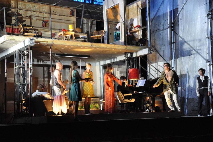 RHEINGOLD, Staatsschauspiel Dresden, Bühne und Kostüme Alissa Kolbusch. ein 2- stöckiges Rohbau- Haus, provisorisch eingerichtet, mit einer Treppe, Geländer und Möbeln. Unten ein Pianist am Flügel und 6 Darsteller,*innen, die singen, oben ein Mann in einer Tonkabine.