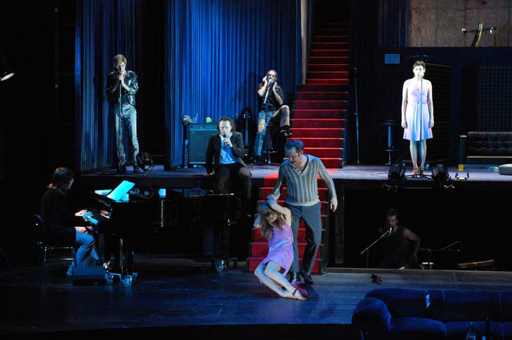LULU nach Wedekind, Staatstheater Hannover, Bühne und Kostüme: Alissa Kolbusch. Ein dreistöckiger blauer Raum mit einer roten langen Treppe. Vier Darsteller machen Musik, zwei stehen an Mikrofonen und singen. Unten Zerrt ein Darsteller eine Darstellerin im Kleid zur Treppe. Überall in den Räumen sind Sessel und Instrumente verteilt.