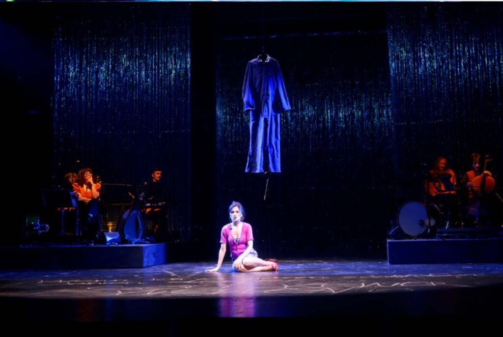 UNSER DEUTSCHLANDMÄRCHEN, Darstellerin sitzt auf einer Theaterbühne unter einem schwebenden Kleidungsstück, während Musiker seitlich vor einem glitzernden Vorhang spielen.