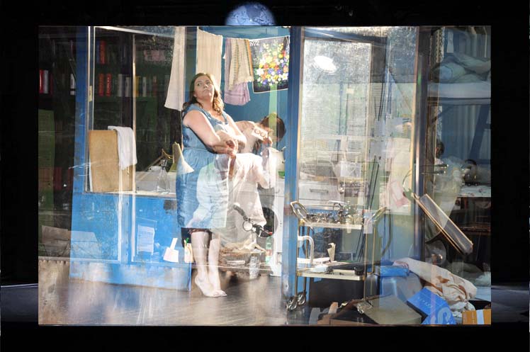 HARMONIA CAELESTIS, Kasino Burgthater Wien, Ausstattung: Alissa Kolbusch.Ein sehr großer herrschaftlicher klassizistischer Raum mit Stuck, Marmorsäulen, einer Bibliothek , Blumenteppich, Stehlampen und antiken Möbeln. Nahaufnahme auf ein blaues Kiosk- Häuschen, wo eine Frau in blauem Kleid sich anlehnt. Im Häuschen Küchenutensilien und eine Wäscheleine mit Handtüchern.