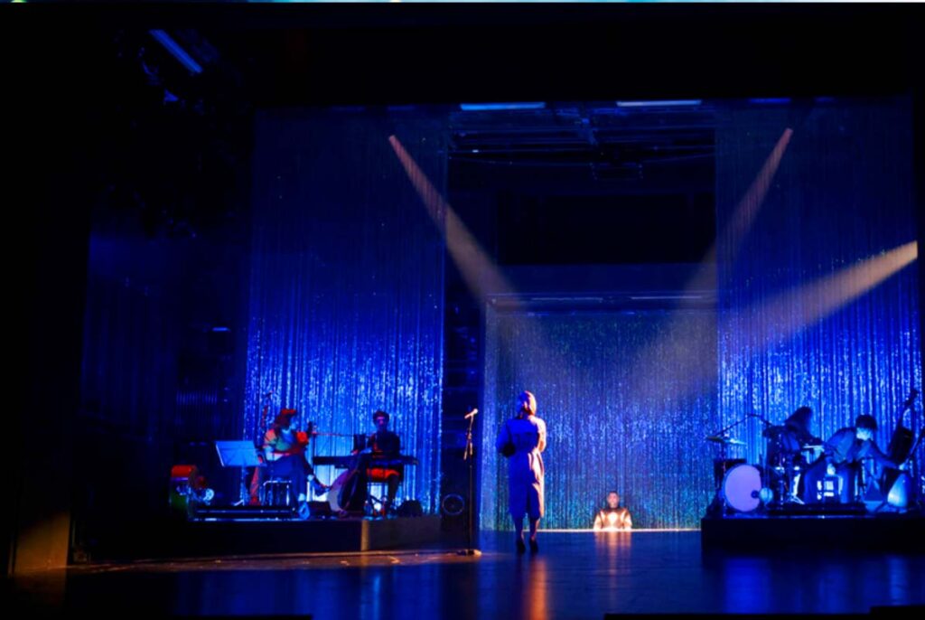 Darstellerin geht über eine blau beleuchtete Theaterbühne vor glitzerndem Vorhang, während Musiker auf beiden Seiten spielen.
