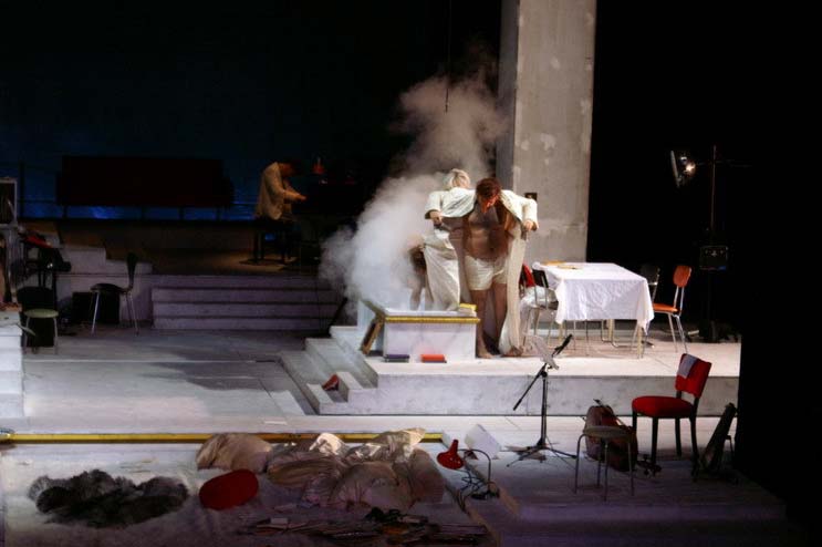 DIE KRÖNUNG DER POPPEA, Bühne und Kostüme: Alissa Kolbusch. Eine große weisse Wohnlandschaft mit einem Podest, auf dem eine Badewanne und ein Esstisch stehen, sowie ein Türrahmen mit Tür. Unten eine Einbuchtung mit Flokati- Teppich, Bettdecken und Leselampe, hinten ein Wohnzimmer mit Bücherregalen, Sesseln und einem E- Piano. Der Mann im langen weißen Mantel steigt in die Badewanne, aus der Nebel emporsteigt . Hinten ein Musiker am Flügel .