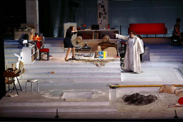 DIE KRÖNUNG DER POPPEA, Bühne und Kostüme: Alissa Kolbusch. Eine große weisse Wohnlandschaft mit einem Podest, auf dem eine Badewanne und ein Esstisch stehen, sowie ein Türrahmen mit Tür. Unten eine Einbuchtung mit Flokati- Teppich, Bettdecken und Leselampe, hinten ein Wohnzimmer mit Bücherregalen, Sesseln und einem E- Piano. Ein Mann steht vorne in einem langen weißen Mantel und streitet mit eienr Frau im Wohnzimmer . ein Musiker am E-piano.