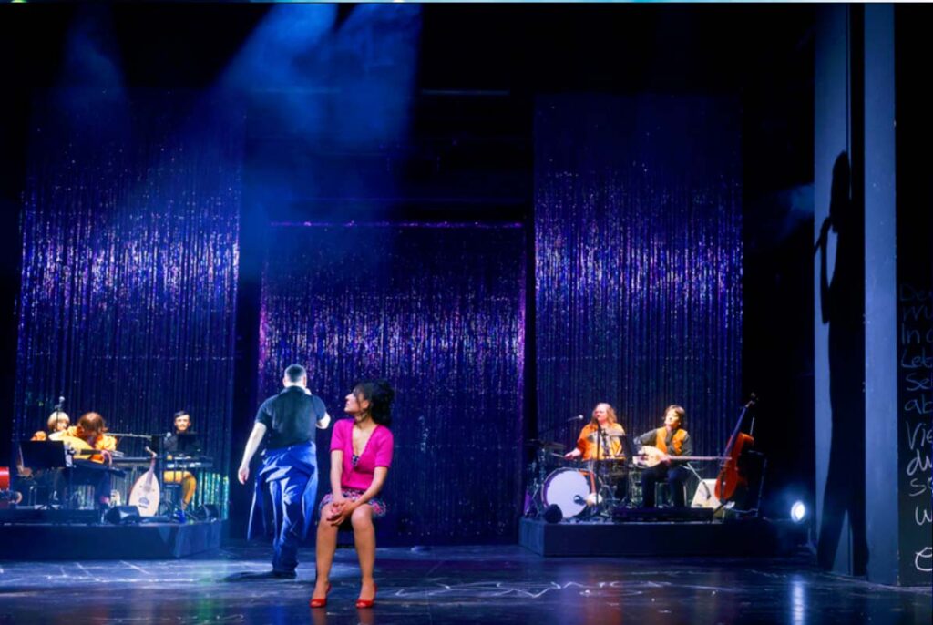 UNSER DEUTSCHLANDMÄRCHEN, Darstellerin sitzt auf einer Theaterbühne vor glitzerndem Vorhang, während ein Darsteller und Musiker im Hintergrund auftreten.