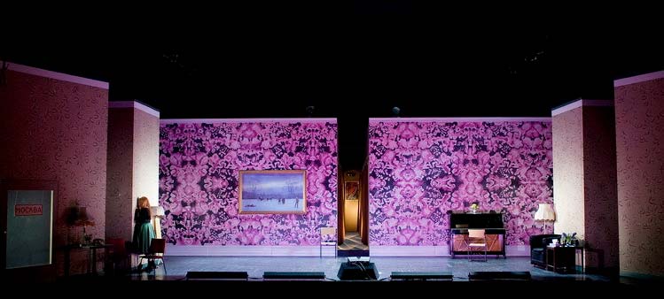 EUGEN ONEGIN, Bühnenbild Alissa Kolbusch. Eine lange Wand mit pinkfarbener Blumentapete , links hängt ein blaues Gemälde an der Wand. In der Mitte teilt sich die Wand und man sieht einen schmalen Korridor. Eine Frau steht links mit dem Gesicht zur Wand und weint.
