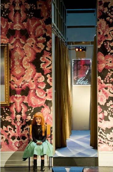 EUGEN ONEGIN, Bühnenbild Alissa Kolbusch. Nahaufnahme auf eine lange Wand mit pinkfarbener Blumentapete , links hängt ein blaues Gemälde an der Wand. In der Mitte teilt sich die Wand und man sieht einen schmalen Korridor, Am ende hängt ein Kinoplakat. Vorne links sitzt ein kleines Mädchen auf einem Schul- Stuhl.
