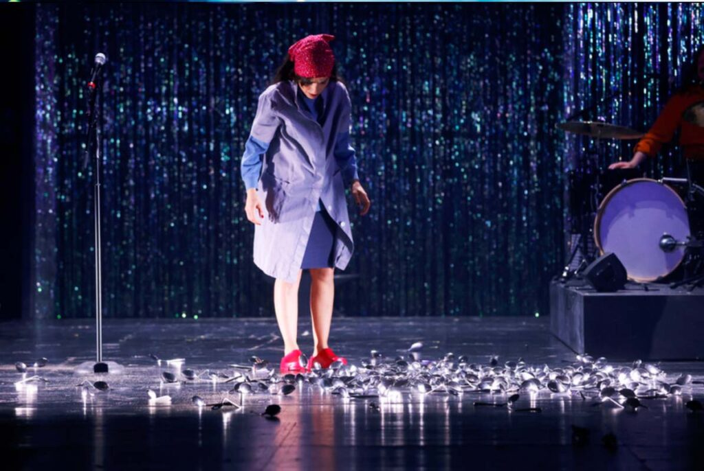 UNSER DEUTSCHLANDMÄRCHEN,Darstellerin auf einer Theaterbühne blickt auf viele verstreute Löffel am Boden, vor einem glitzernden Vorhang und neben einem Mikrofonständer.