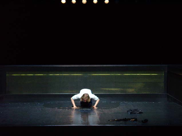 DAS MISSVERSTÄNDNIS, Deutsches Theater, Bühne: Alissa Kolbusch. Eine Frau kniet in der Mitte der Bühne in einer Wasserpfütze , schaut und spricht in das Wasser auf dem Boden.