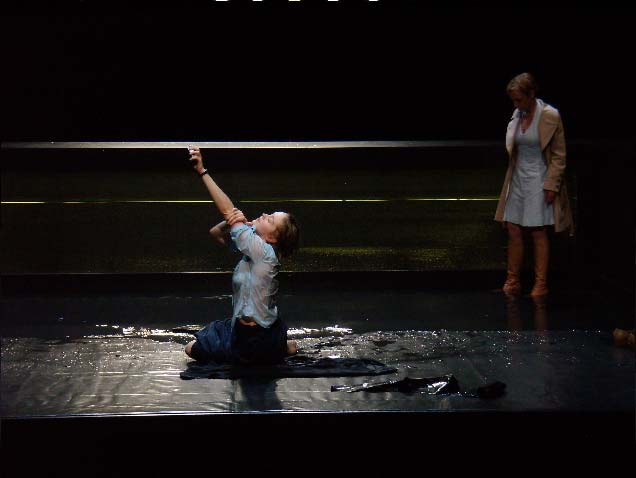 DAS MISSVERSTÄNDNIS, Deutsches Theater, Bühne: Alissa Kolbusch. Eine Frau sitzt in der Mitte der Bühne in einer Wasserpfütze , die andere Frau steht hinter ihr mit den Stiefeln im Wasser.