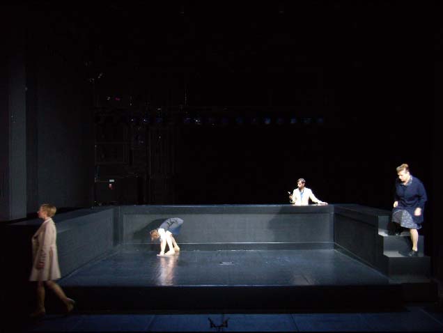 DAS MISSVERSTÄNDNIS, Deutsches Theater, Bühne: Alissa Kolbusch. Eine Frau steht gebeugt in der Mitte der schwarzen Bühne in einer Wasserpfütze, ein Mann steht hinter ihr hinter dem Steg , die anderen 2 Frauen bewegen sich links vorne und rechts steigt eine die Stufen vom Steg herunter.