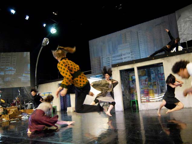 MEGALOPOLIS, Schaubühne Berlin, Bühnenbild Alissa Kolbusch. Zwei flache Häuser, die Dächer mit einem Steg verbunden. Links eine große Straßenlaterne und Kartons vor dem Haus. Hinten eine gemalte Großstadt. Drei Tänzerinnen springen, 4 andere Tänzer*innen tanzen auf dem Boden und auf dem dach des Hauses.