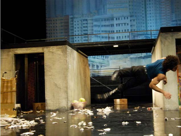 MEGALOPOLIS, Schaubühne Berlin, Bühnenbild Alissa Kolbusch. Zwei flache Häuser, die Dächer mit einem Steg verbunden. Links eine große Straßenlaterne und Kartons vor dem Haus. Hinten eine gemalte Großstadt. Ein Polizist springt und liegt quer in der Luft .