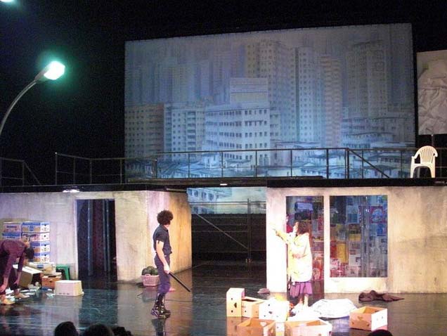 MEGALOPOLIS, Schaubühne Berlin, Bühnenbild Alissa Kolbusch. Zwei flache Häuser, die Dächer mit einem Steg verbunden. Links eine große Straßenlaterne und Kartons vor dem Haus. Hinten eine gemalte Großstadt. Eine Frau gestikuliert und ein Polizist schaut nach hinten.