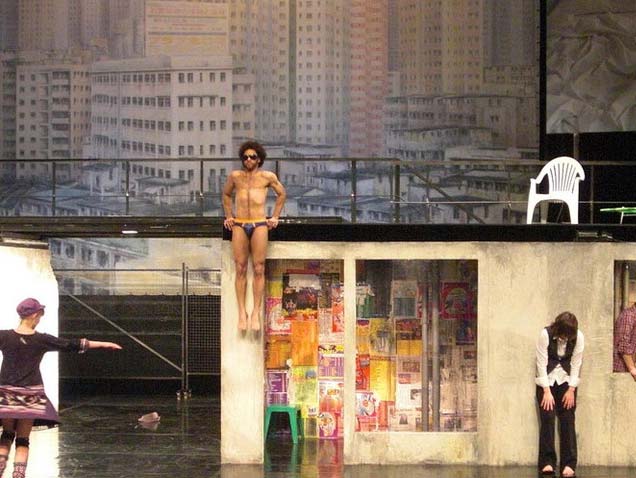MEGALOPOLIS, Schaubühne Berlin, Bühnenbild Alissa Kolbusch. Ein flaches Haus, an der Vorderkante macht ein Mann in einer badehose Klimmzüge, Schlagzeuger. Vor dem Haus eine Frau, die sich nach vorn beugt , eine andere breitet die Arme aus. Hinten eine gemalte Großstadt.