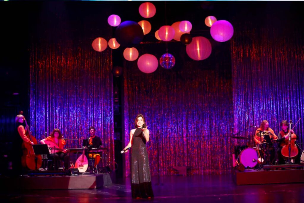 UNSER DEUTSCHLANDMÄRCHEN,Sängerin steht auf einer Theaterbühne vor glitzerndem Vorhang und bunten Lampen, während Musiker auf beiden Seiten spielen.