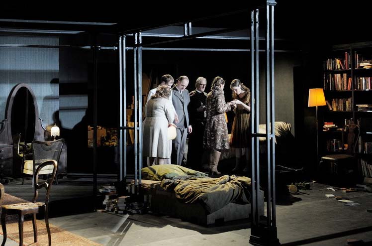 DAS WOHLTEMPERIERTE KLAVIER, Schaubühne Berlin/MC93 Bobigny, Bühnenbild Alissa Kolbusch. Ein dunkles breites Wohnzimmer mit antiken Stühlen und einem Flügel der ohne Beine auf dem Teppich liegt. Sechs Darsteller stehen in einer Gruppe im Raum rechts neben dem Wohnzimmer, in dem eine Bibliothek ist und ein Bett steht.