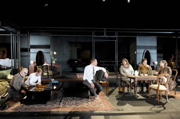 DAS WOHLTEMPERIERTE KLAVIER, Schaubühne Berlin/MC93 Bobigny, Bühnenbild Alissa Kolbusch. Ein dunkles breites Wohnzimmer mit antiken Stühlen und einem Flügel der ohne Beine auf dem Teppich liegt. Vier Darsteller sitzen auf dem Stühlen in der Mitte und sprechen miteinander, 2 Männer knien links und spiele auf dem Flügel.