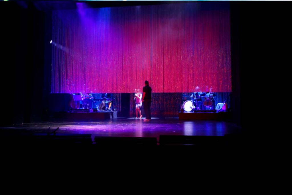 UNSER DEUTSCHLANDMÄRCHEN, Theaterszene mit zwei Darsteller:innen vor einem rot schimmernden Vorhang, während Musiker auf beiden Seiten der Bühne spielen.