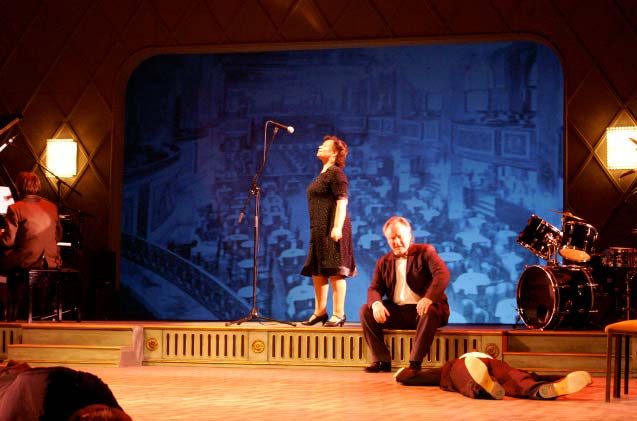 CAFE VATERLAND, Maxim Gorki Theater, Bühne und Kostüme: Alissa Kolbusch. Ein Ballsaal mit einem Flügel. die ältere Dame steht auf der Bühne und versucht in ein Mikrofon zu singen, das zu hoch für sie ist, der ältere Herr sitzt an der Rampe. Hinten auf einer Leinwand ein Ballsaal.