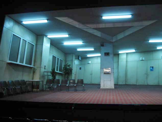 CAFE UMBERTO, Theater Bremen, Bühne und Kostüme: Alissa Kolbusch. Ein ungemütlicher "Ämter"-Raum mit einer schrägen Decke mit Neonleuchten und einer Säule in der Mitte. 2 Fenster und eine Sitzecke links.