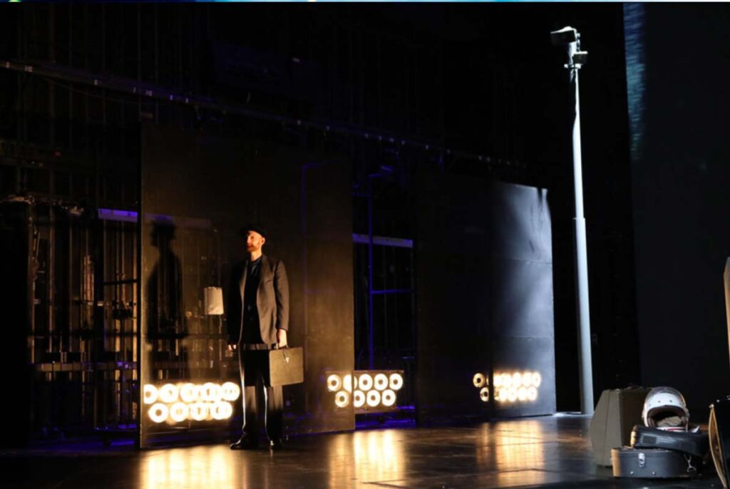 DER REISENDE, Darsteller mit Koffer steht auf einer Theaterbühne neben einem hohen Laternenmast vor einer dunklen Bühnenkulisse mit Scheinwerfern.